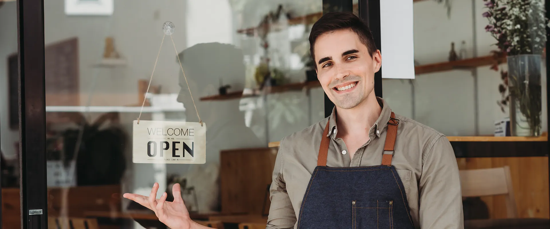 Un commerçant accueille ses clients devant sa boutique, pancarte « Open »
